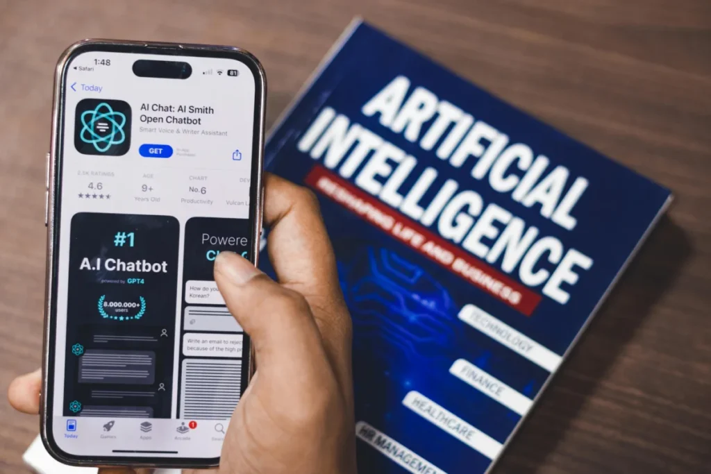 Person holding a smartphone displaying an AI chatbot app on the App Store, with a book titled “Artificial Intelligence” on a table in the background.
