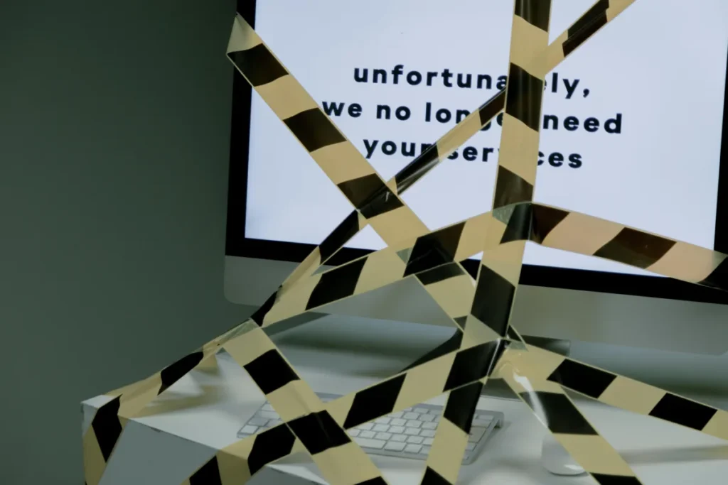 A computer monitor covered with black and yellow caution tape displaying a message that reads, “unfortunately, we no longer need your services,” symbolizing job loss or termination.