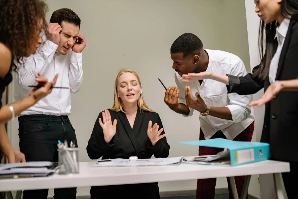 : A group of five coworkers stand around a desk during a heated discussion. One woman sitting at the desk has her eyes closed and hands raised, appearing to stay calm, while others around her gesture intensely and appear frustrated or upset.
