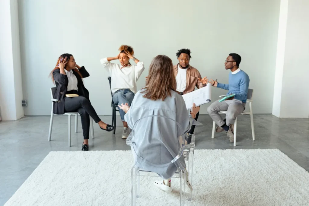 A group of five people sit in a circle in a bright room during a discussion or therapy session. One person, with their back to the camera, appears to be leading the session and holding papers, while the others engage in conversation and expressive gestures
