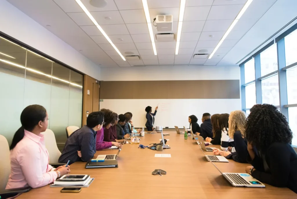 Team of professionals in a conference room watching a presenter write on a whiteboard during a business meeting.