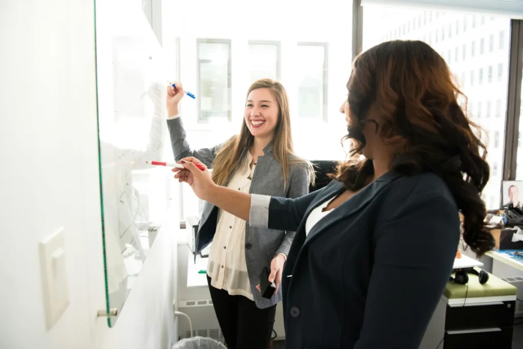 Two businesswomen smiling and writing on a whiteboard together during a brainstorming session in a bright office
