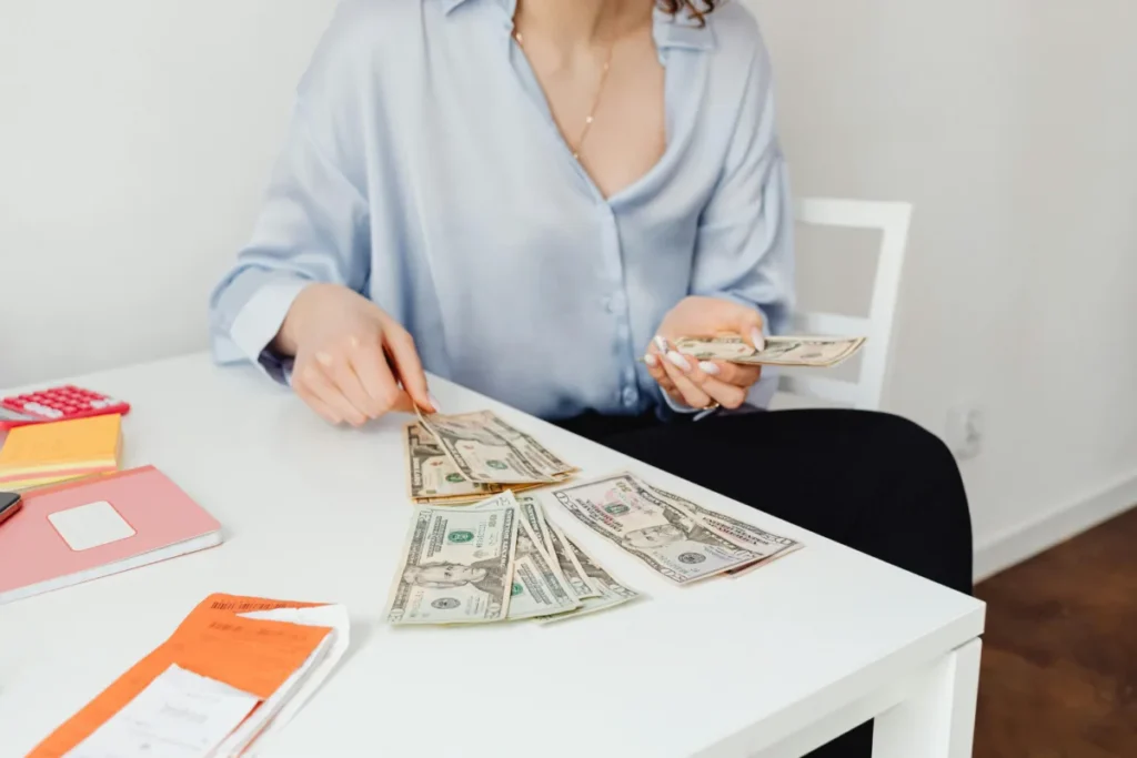 Person sitting at a desk counting U.S. dollar bills with notebooks, receipts, and a calculator nearby, representing budgeting or financial planning