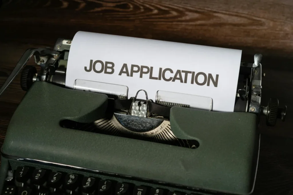 A close-up image of a vintage green typewriter with a sheet of paper inserted that reads "JOB APPLICATION" in bold letters. The background features a wooden surface.