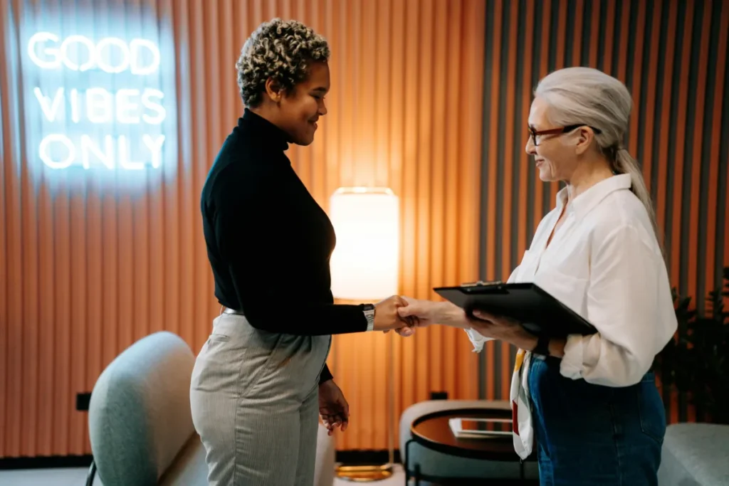 Two women smiling and shaking hands in a modern office setting. One is holding a clipboard, and a neon sign on the wall behind them reads “GOOD VIBES ONLY.” The atmosphere appears friendly and professional