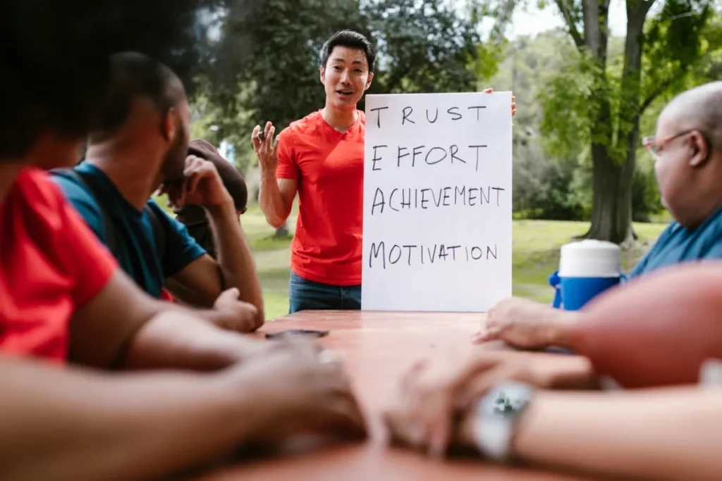 A person standing outdoors giving a presentation to a small group seated at a table. The presenter is holding a large white poster board with the words "TRUST," "EFFORT," "ACHIEVEMENT," and "MOTIVATION" written on it.