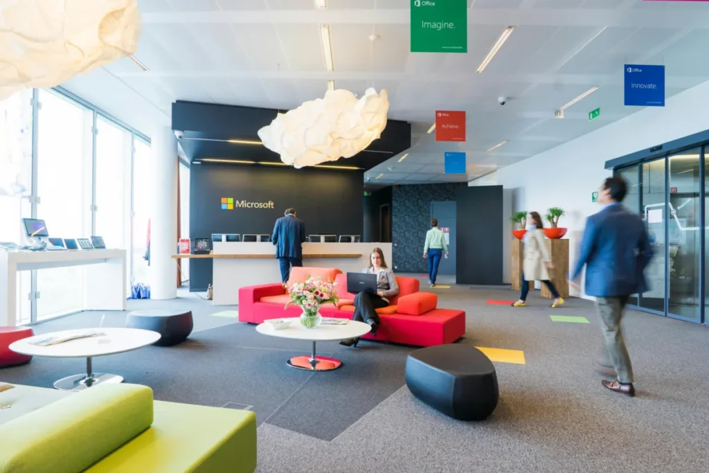 A modern office lobby with colorful furniture, featuring the Microsoft logo on a dark wall behind a reception desk. Several people are walking or working, and a woman is sitting on a red sofa using a laptop.