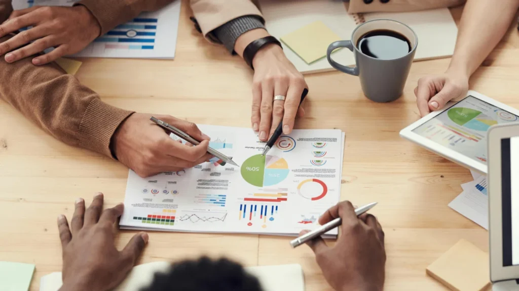 Group of people reviewing business charts and graphs on paper and a tablet during a meeting, with coffee and pens on the table.