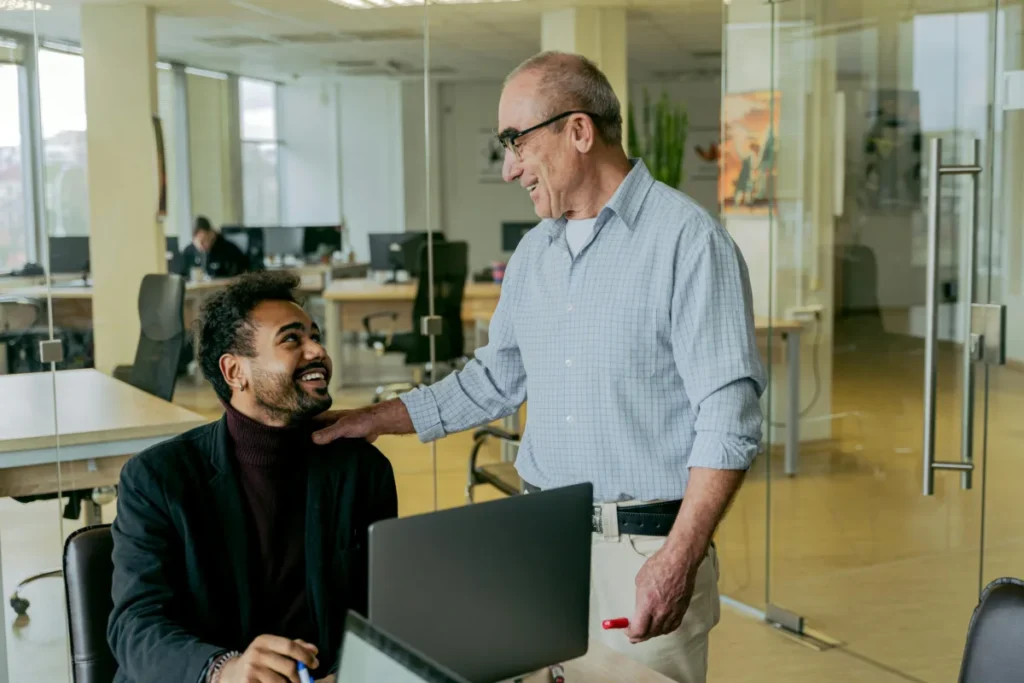 Smiling older man standing and talking with a younger colleague sitting at a desk in a modern office environment.