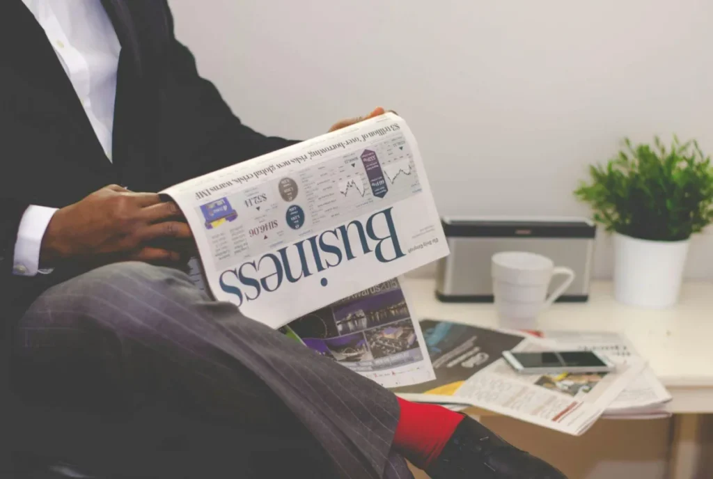 A person in a business suit and red socks is sitting and reading a newspaper with the headline “Business.”