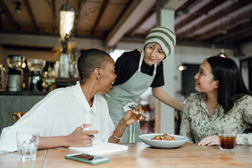 In a cozy café, a server checks in with two women; one gestures with a pen over a notebook while the other smiles, suggesting guidance and collaborative dialogue—an informal moment of leadership development in action.