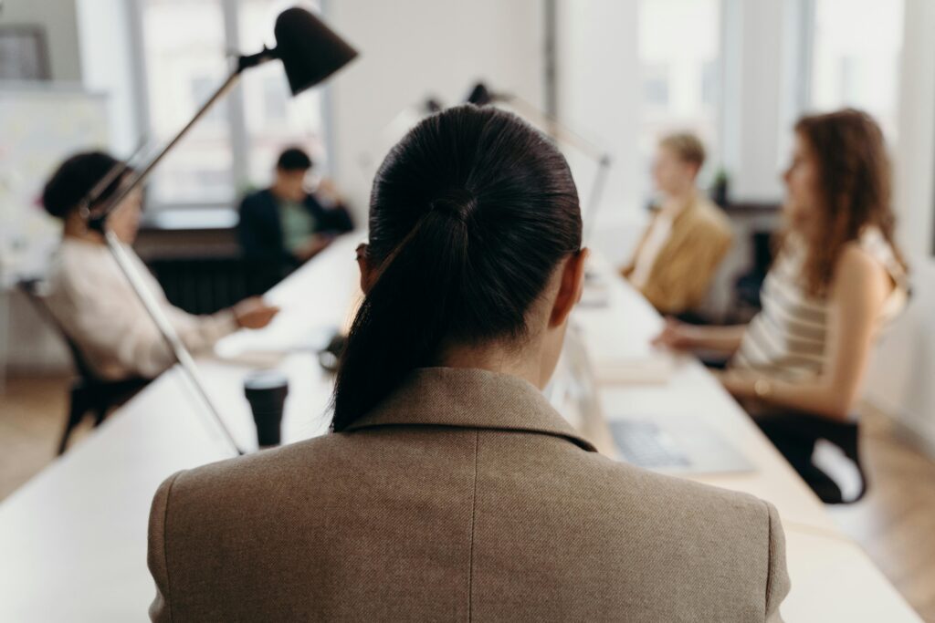 An office setting reflecting leadership weaknesses, showing a woman viewed from behind as she faces her team across a long desk, symbolizing distance from the group.