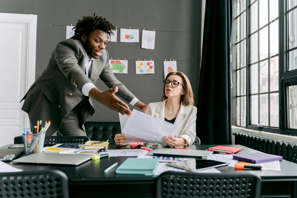 A man in a suit leans in and gestures aggressively at a seated coworker who looks frustrated, depicting a toxic culture at work characterized by intimidation and disrespect.