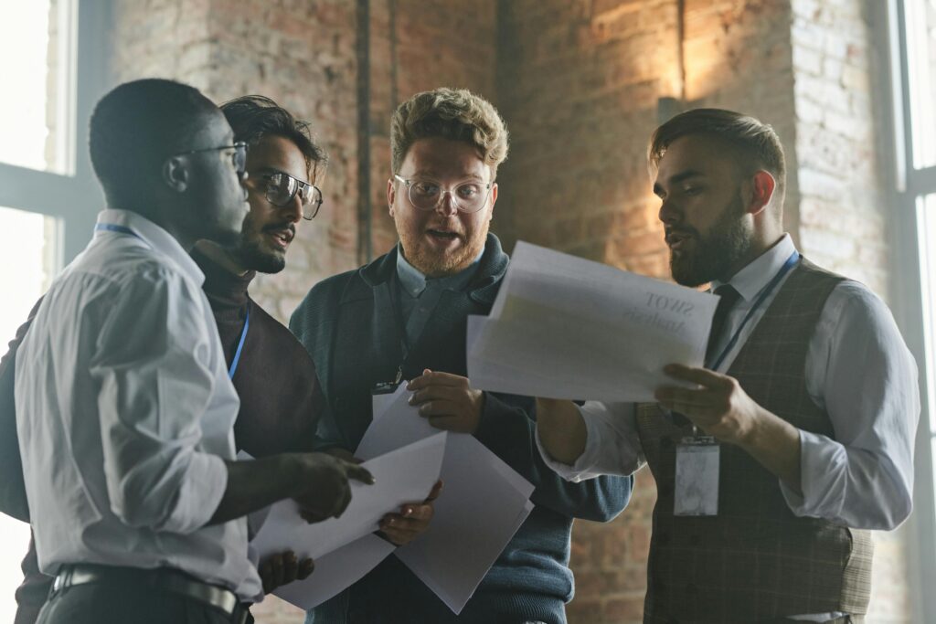 Four colleagues huddle tensely over printed sheets near a window, their anxious expressions signaling a toxic environment at work with pressure and conflict.