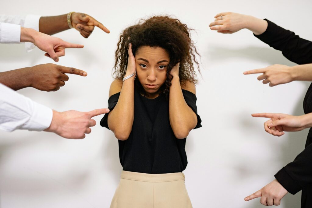 A distressed woman covers her ears as multiple coworkers’ hands point at her from all sides, visualizing a toxic workplace culture of blame and hostility.
