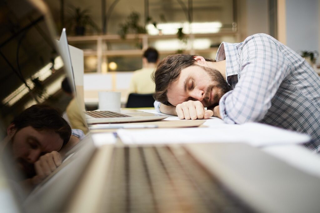 A bearded employee naps on a desk beside an open laptop and scattered papers, capturing a toxic workplace environment marked by exhaustion and burnout.