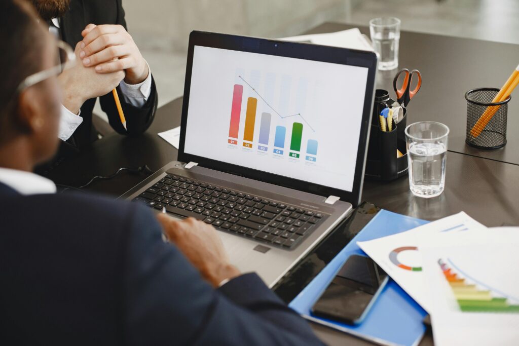 Over-the-shoulder view of a laptop displaying a multicolored bar chart with a downward trend line; documents, phone, and glasses of water on the table emphasize a professional review of business analytics.