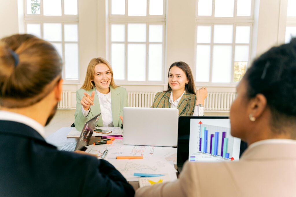 Four colleagues sit around a table with laptops and printouts, discussing results as a screen shows a bar chart; bright windows and casual gestures suggest a collaborative meeting focused on business data.