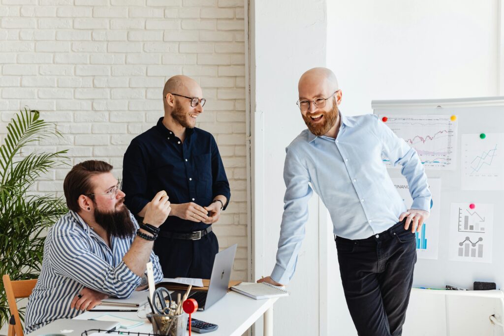Three male colleagues in a light, modern workspace laugh and discuss insights next to a whiteboard covered with line graphs and charts, illustrating teamwork and business intelligence collaboration.