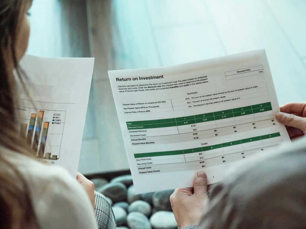 Close-up of two people reviewing printed ROI spreadsheets; one sheet shows discount factors and cost/benefit tables while the other displays a multicolored bar chart, conveying financial data analysis in a modern office setting.